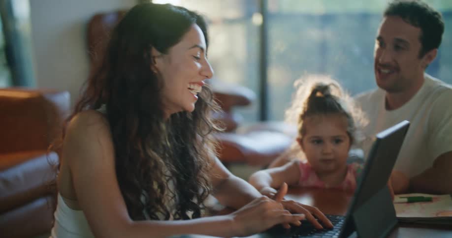 A man sits with his young daughter on his lap as he talks with his wife who is laughing while using her laptop