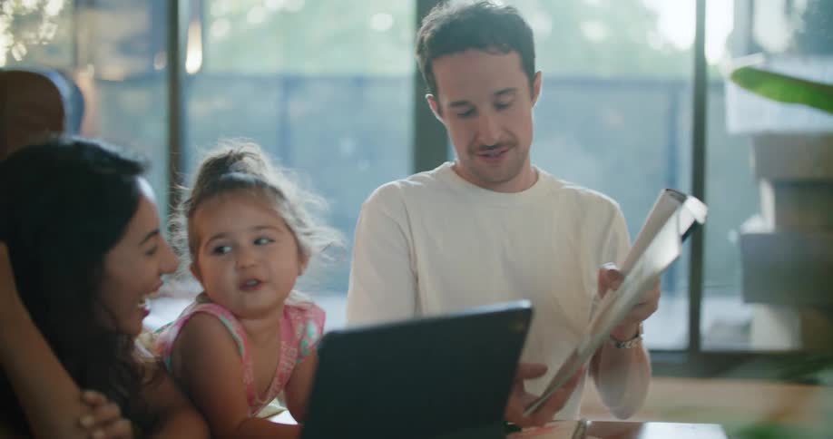 Toddler girl leans against coffee table between father with mustache white t shirt and mother with wavy hair and sleeveless white shirttalking and reading magazine in living room at home
