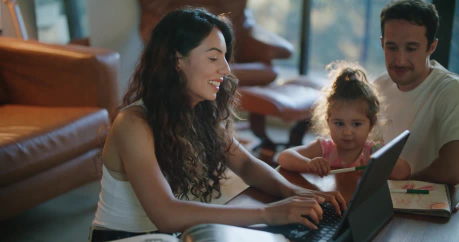 Father with mustache white t shirt holding toddler girl making a drawing in notebook next to mother with wavy hair using tablet at coffee table