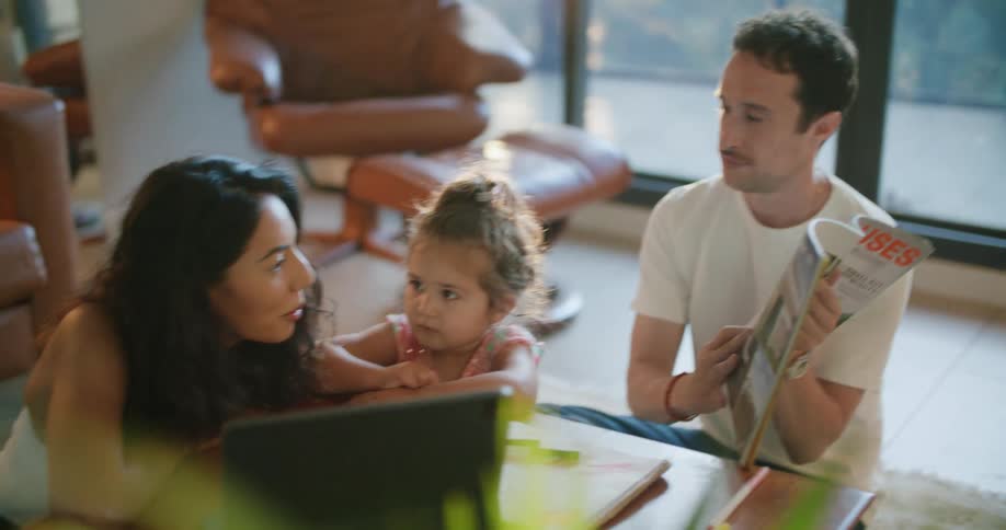 Father with mustache and white t shirt shows magazine to toddler girl and mother with wavy hair using tablet at coffee table