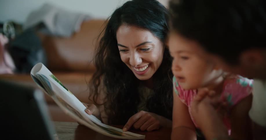 Toddler girl leans against coffee table reading magazine between father with mustache white t shirt and mother with wavy hair and sleeveless white shirt