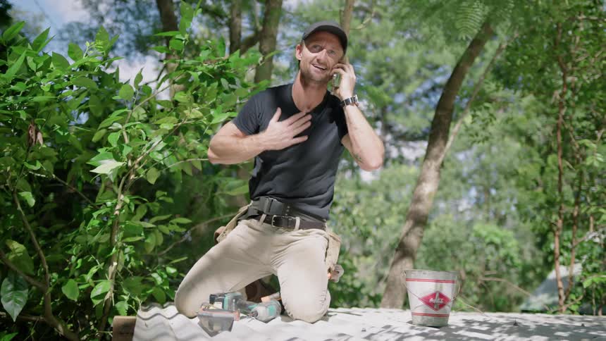 A man talks on his smartphone while kneeling on top of his roof doing yardwork