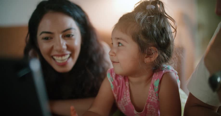 Mother with wavy hair and toddler girl watch tablet next to father wearing white t shirt and wristwatch in living room at home