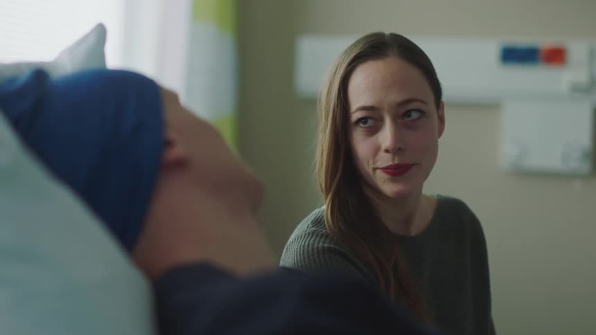 A woman is sitting down talking with a man lying in a hospital bed