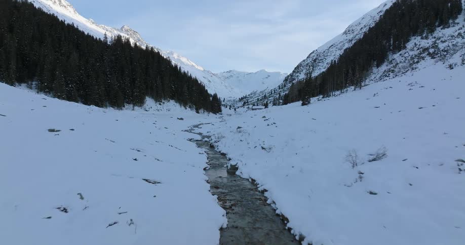 A stream runs through a snow covered valley with a forest of trees along the mountainside