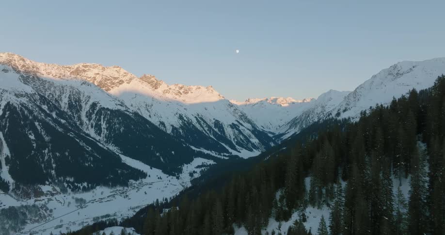 Aerial view over a forest of trees on a snow covered mountain and the moon over a snow covered mountain range in the distance