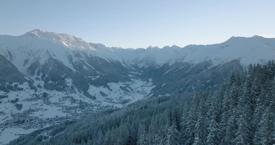 The sun shines on snow covered mountains and an arial view over a forest of snow covered trees along the side of a mountain