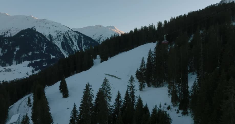 A cable car moves down a snow covered mountain and over trees as the sun shines on snow covered mountains in the distance