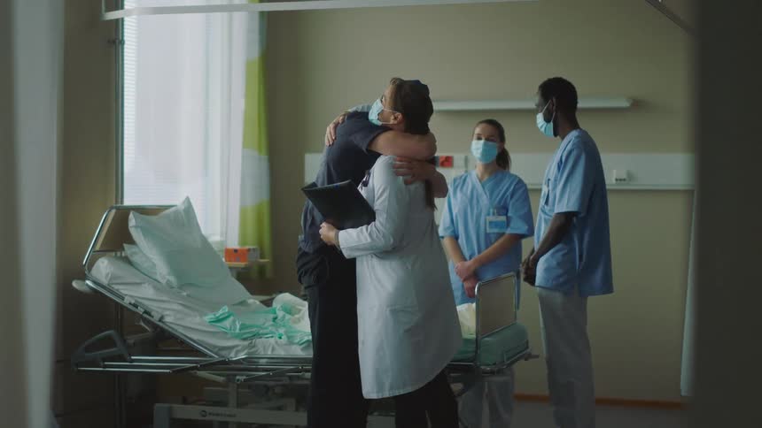 A patient in a hospital bed gets up and hugs a doctor as a woman and two nurses watch