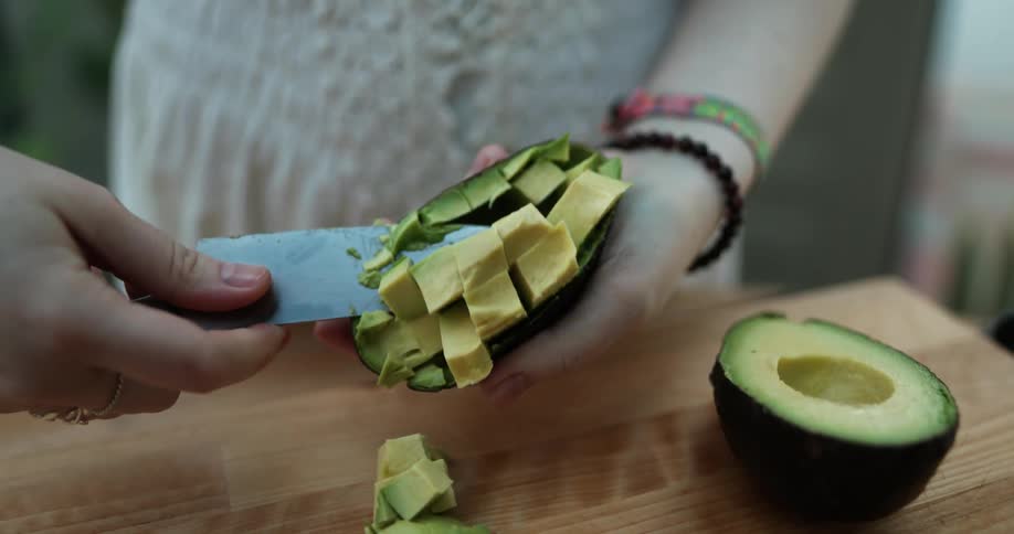 Woman cubes avocados with a knife and dumps pulp onto wooden chopping board