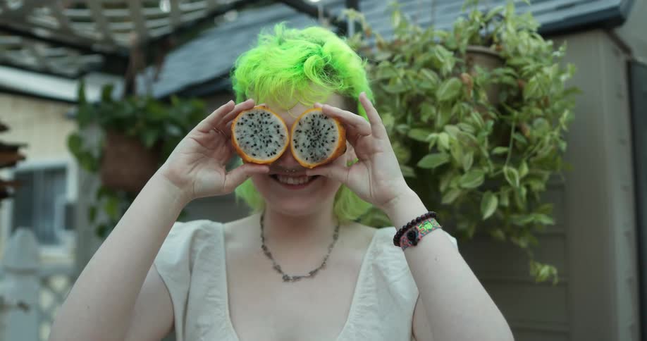 Woman with day glow green hair holds pieces of fruit in front of her eyes as she smiles