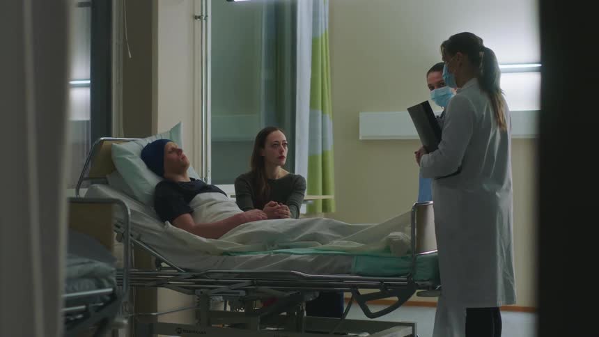 A woman lifts her head and looks at the doctor and nurse while still holding her husbands hand