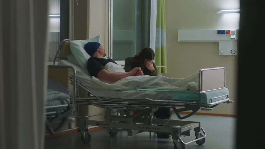 Man laying in a hospital bed holding hands with a crying woman sitting at his side