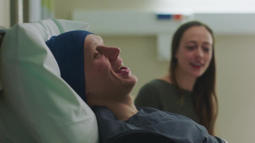 A woman sits next to a mans hospital bed and she shakes her body and they both laugh