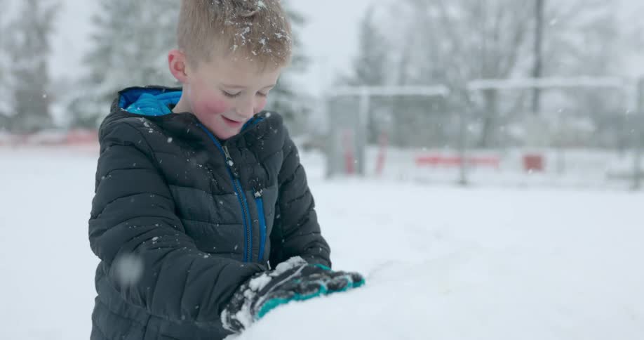 Boy rolls large snowball in snow covered park as it snows