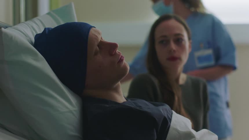 A man drinks a glass of water after taking his medicine then lies back exhausted as his wife sits anxiously