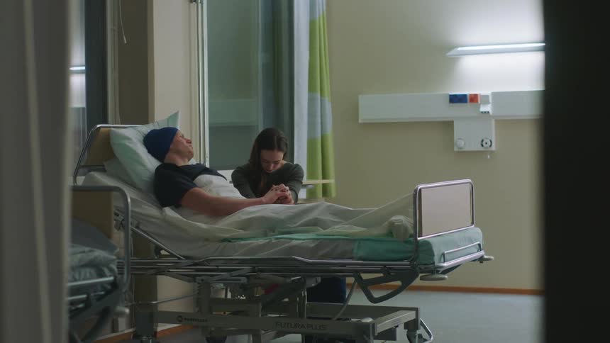 A man lies in a hospital and a woman sits holding his hand and putting her head down as a nurse and doctor leave the bedside