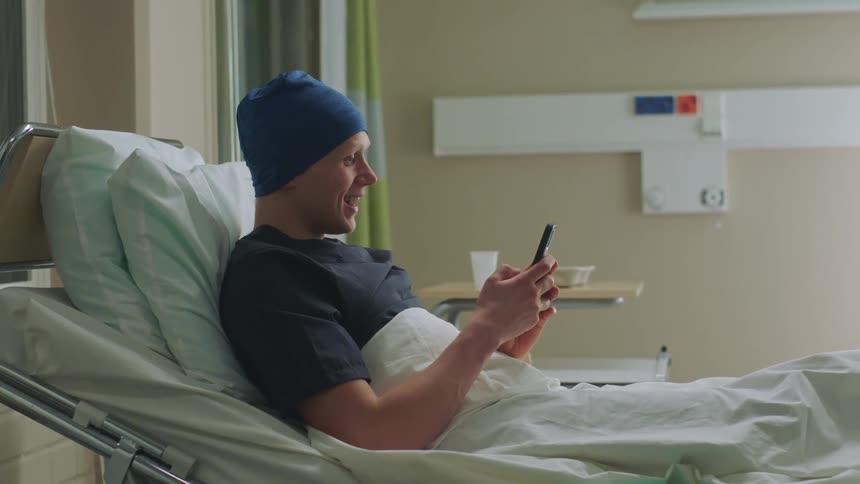 A patient lying in a hospital bed picks up his cell phone and smiles and waves as he video chats