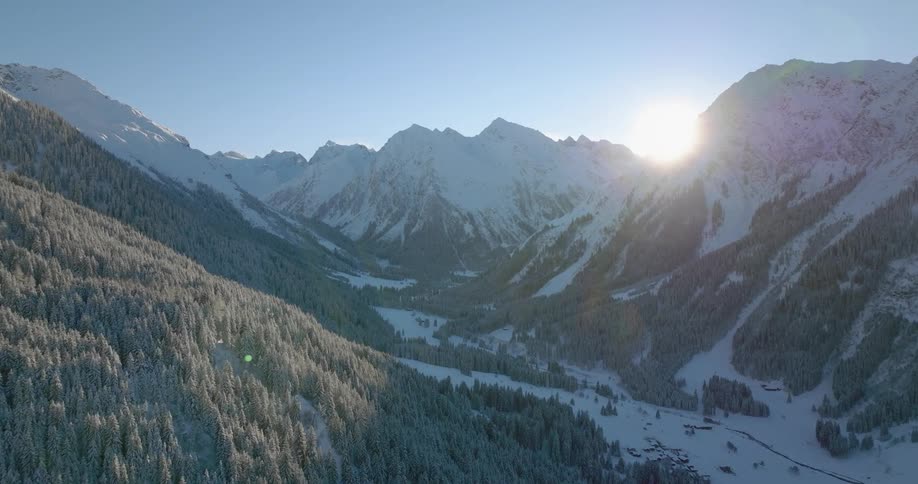 The sun shines brightly over a snow covered mountain range with a forest of snow covered trees along the mountainside