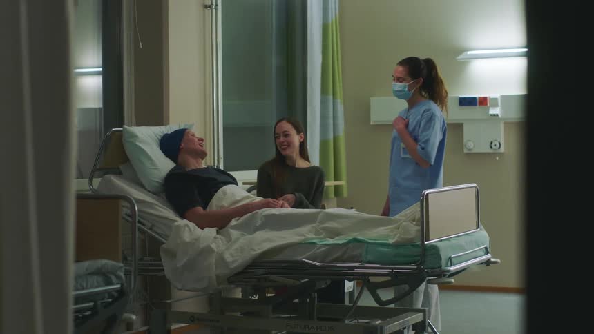 A man lies in a hospital bed and laughs with a woman sitting beside him and a nurse