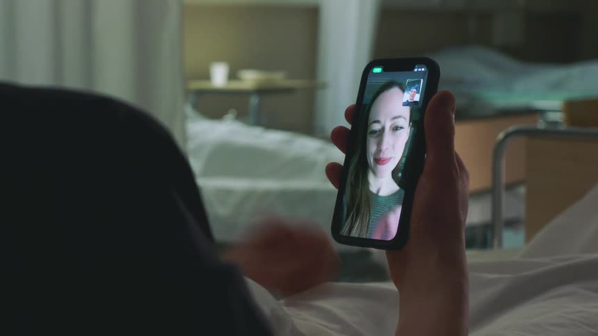 Male patient lying in bed makes a video call on smartphone in hospital room