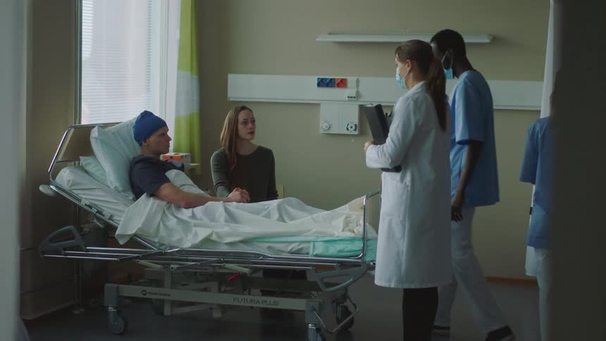 A woman sits next to a mans hospital bed and holds hand and two nurses and a doctor walk in the room and the doctor speaks to them