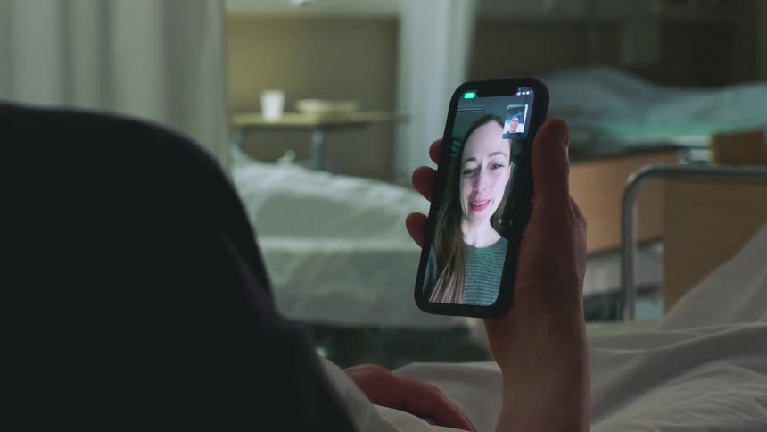 Patient lies in hospital bed waving at and talking with woman smiling and laughing on mobile phone video
