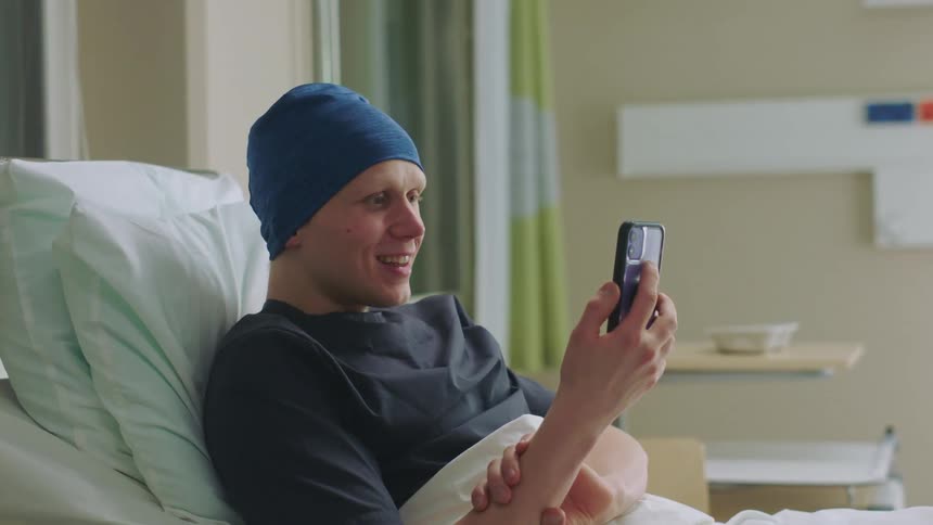 White man lying down at a hospital bed using his smartphone to video call someone