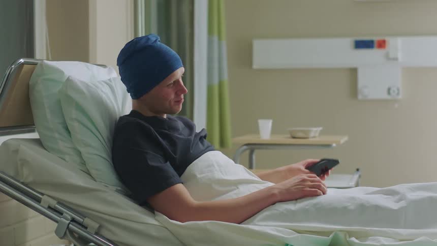A patient lying in a hospital bed waves and talks as he video chats on a cell phone