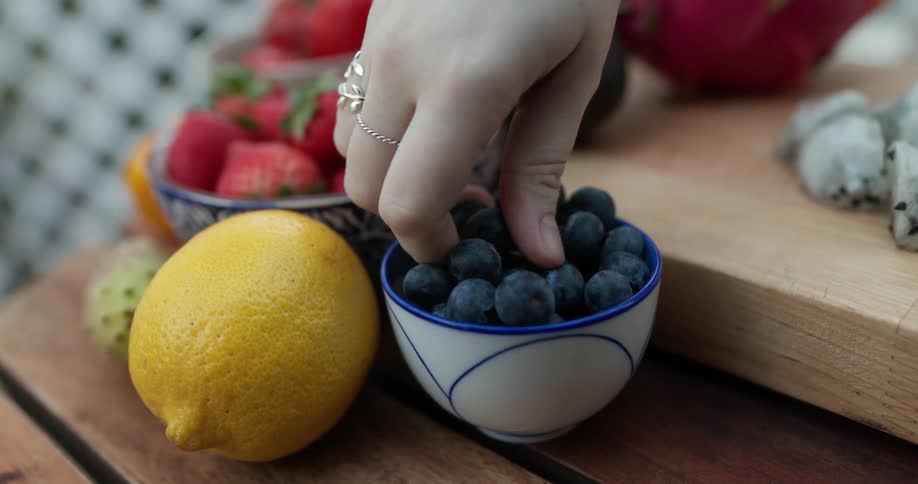 Bowls of fruit are on a table and someone reaches in and takes some blueberries