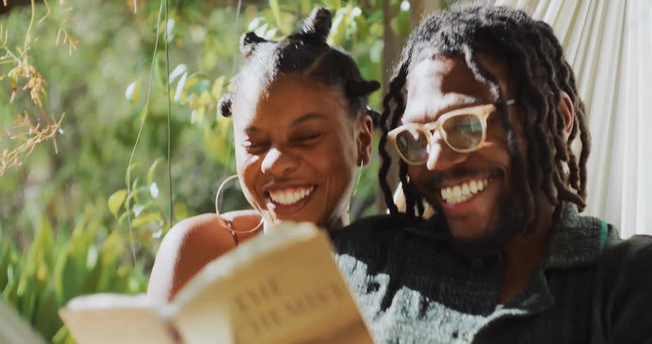 Black man lying down at a hammock with his girlfriend sitting next to him while both are laughing and reading a book together