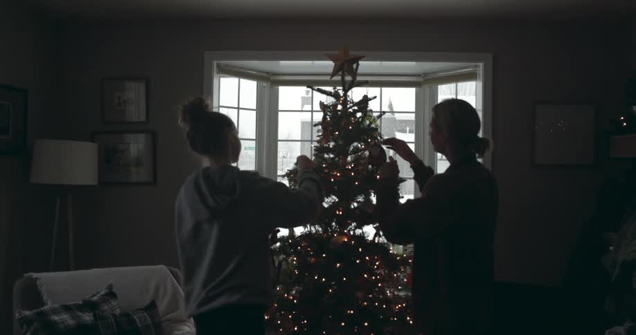 Mother and daughter decorate christmas tree as snow falls outside of bay window