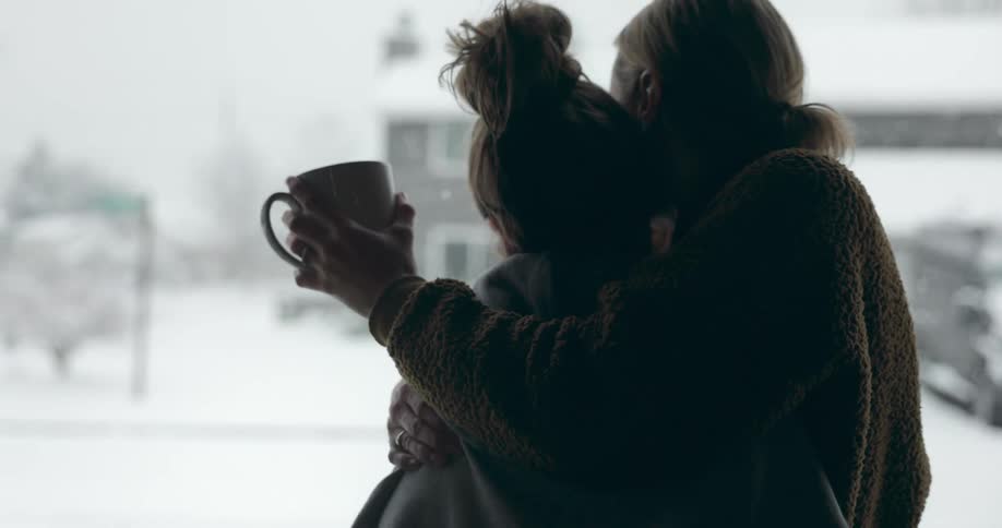 Blonde woman with brown sweater cuddles girl with gray hoodie holding cup looking out the window while snowing