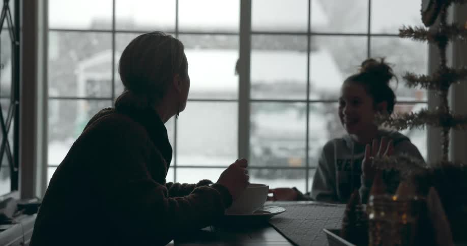 Girl sits at table talking with another person as they eat and snow falls outdoors