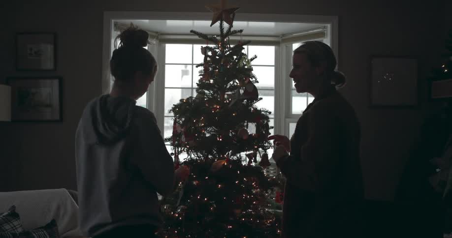 Mother and daughter decorate tree as snow falls outside window