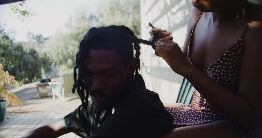 Woman plaits mans hair as they sit on porch near house wall