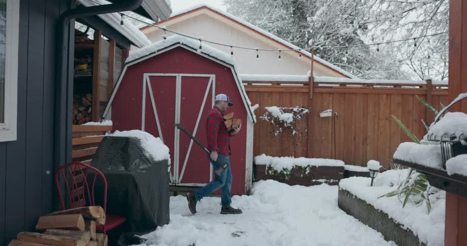 A man is walking outside in his yard in the snow carrying and ax in one hand and cut firewood in the other