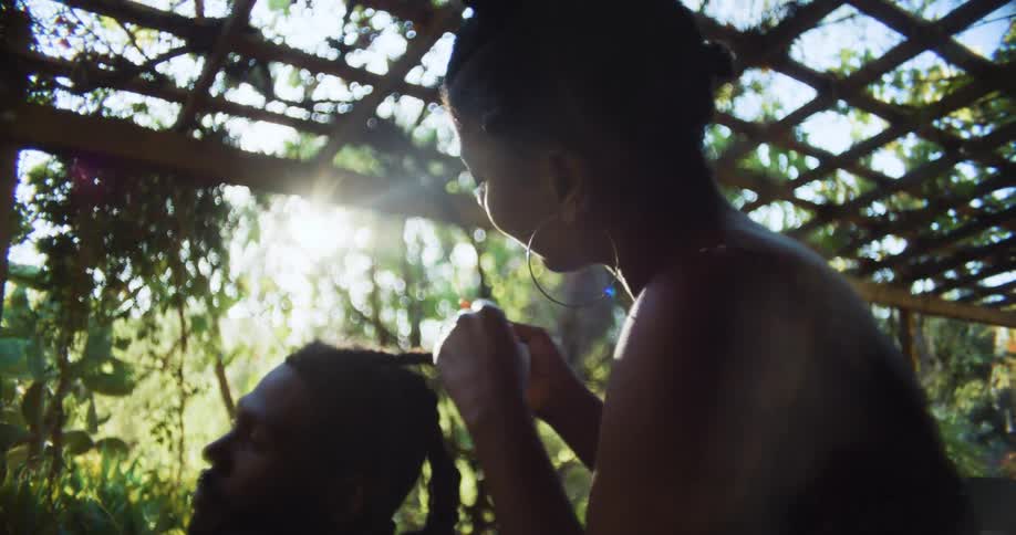 A woman braids a mans hair as they sit under a pergola and she talks to him