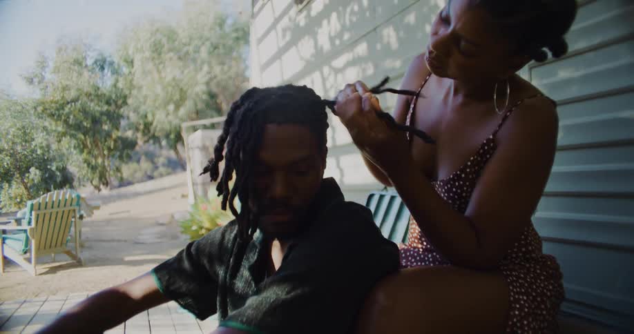Woman plaits mans hair as they sit near mobile home under a wooden overhang
