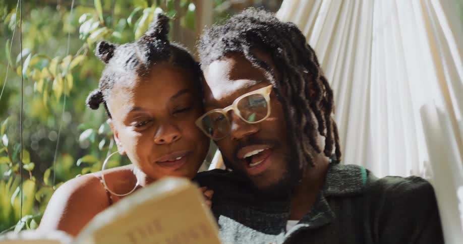 A couple laugh and smile together as a man reads aloud while swaying in a hammock