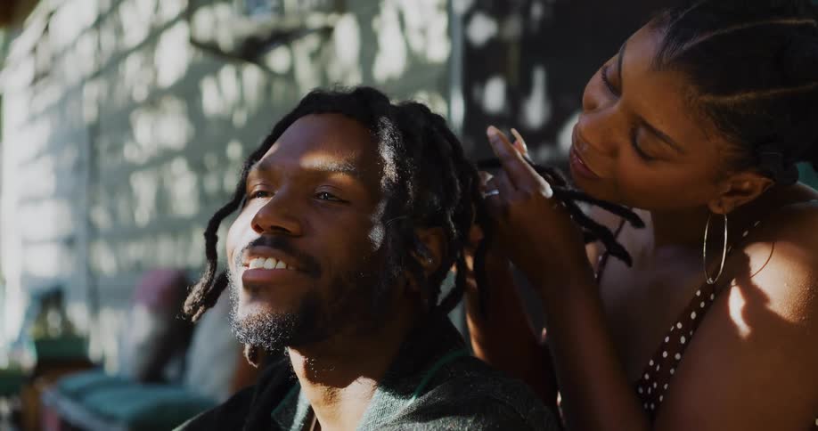 A woman sits on a porch and braids a mans hair as he talks to her