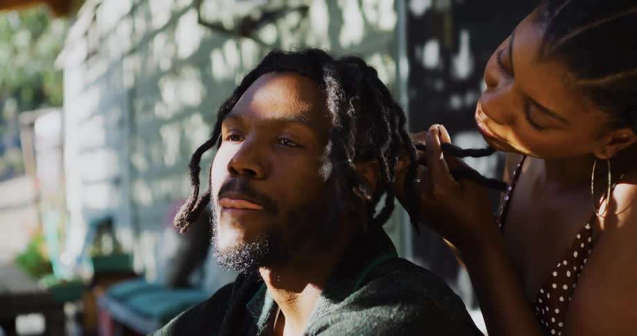 Woman braids the locks of his husband sitting in the porch at home
