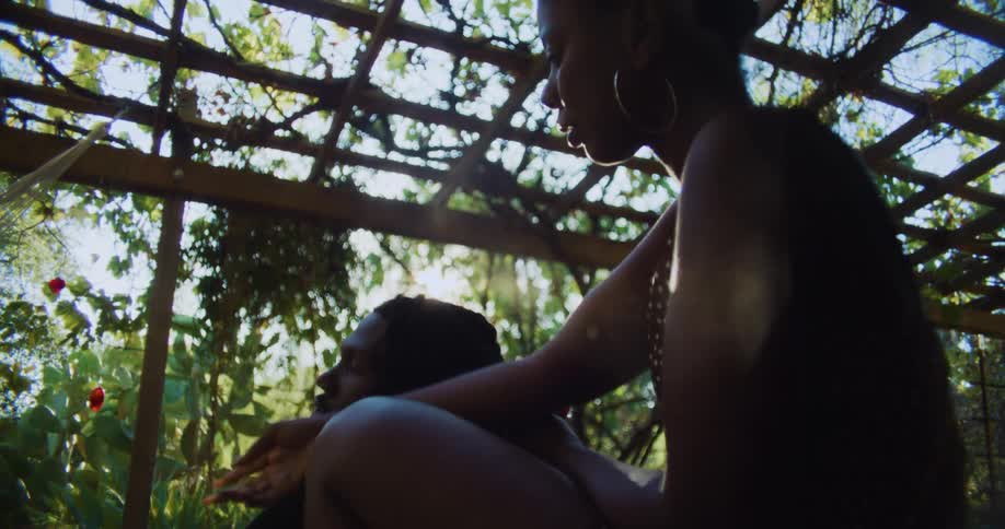A man and woman sit under a pergola and he hands her an elastic as she braids his hair