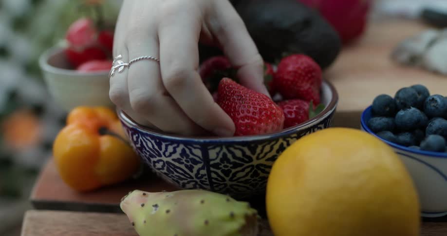 A woman picks a strawberry up out of a bowl on a table with fruits and vegetables on it