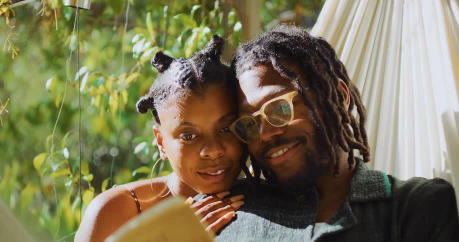 A woman cuddles up to a man as he reads to her in a swaying hammock then they both laugh