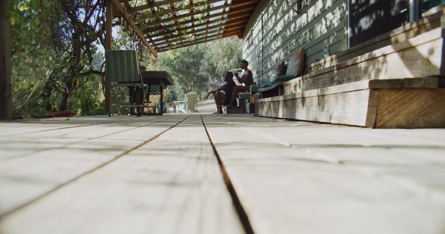 A man and woman sit on a porch and she braids his hair