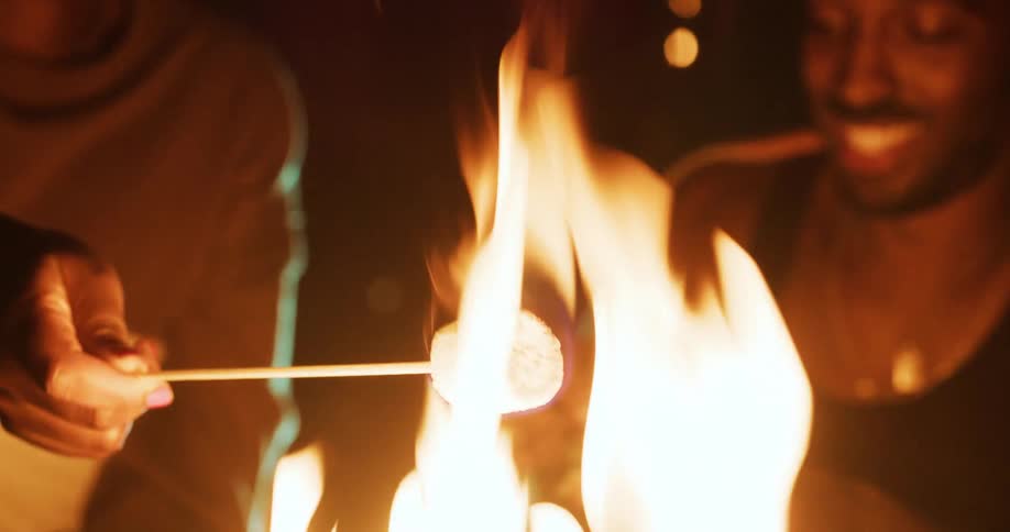 Two black men talking and laughing while sitting next to a bonfire roasting a marshmallow