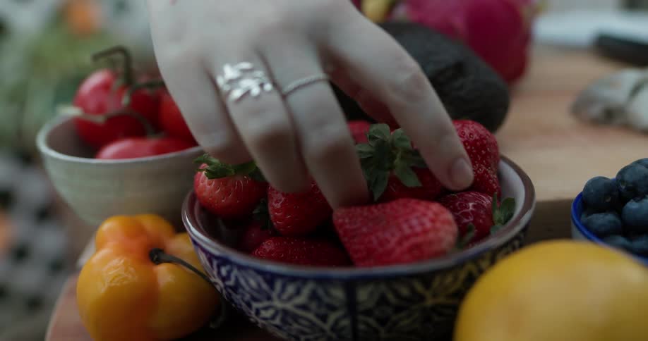 There are bowls of fruit and vegetables on a table and someone picks up a strawberry