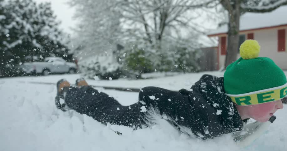 A boy is carrying a sled as he runs down a snow covered street and he slides across the snow riding on his chest