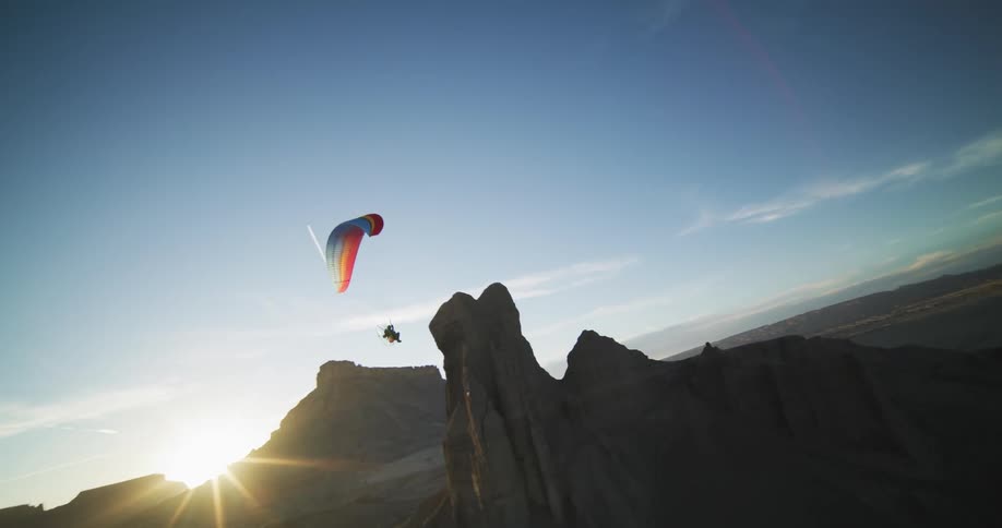 A hang glider flies over rock formations in the desert as the sun shines brightly on the horizon